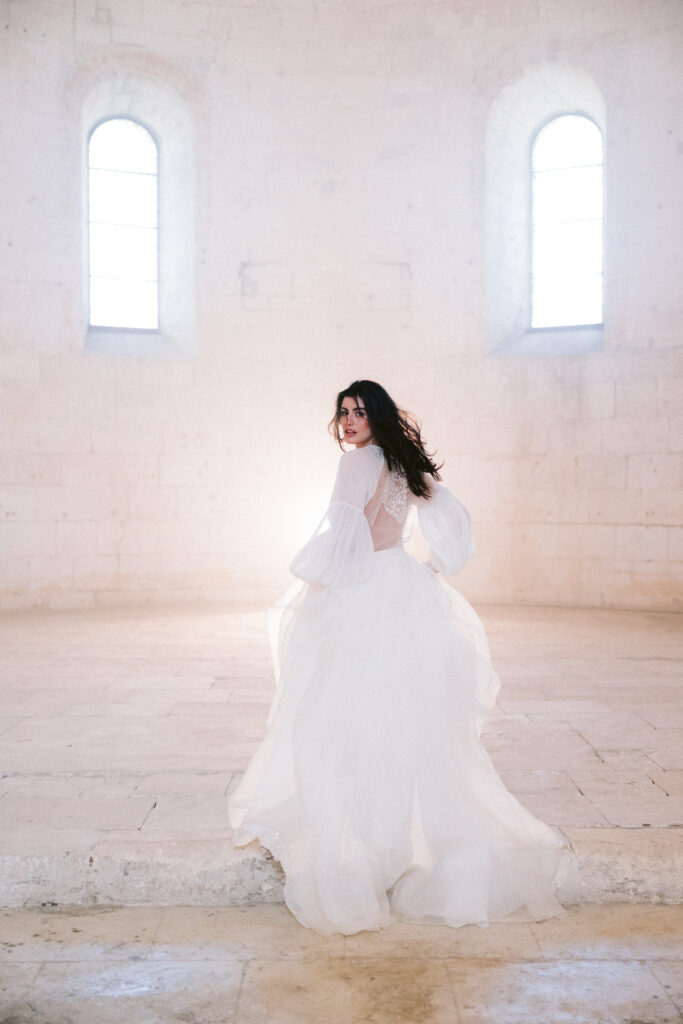 Une magnifique mariée se tourne dans une salle élégante, illuminée par des fenêtres hautes, incarnant le rêve d'un mariage romantique. By Mademoiselle C, wedding planner en Provence, Côte d'Azur et Corse, crée des cérémonies luxueuses sur mesure dans des lieux d'exception. Une célébration grandiose pour un événement unique, dans un cadre sophistiqué et enchanteur.