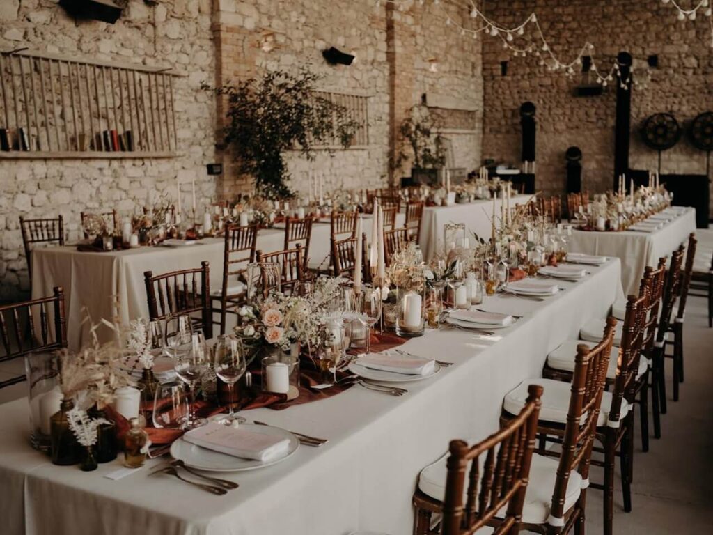 Une élégante salle de réception en pierre, décorée pour une célébration de luxe, mettant en avant le savoir-faire de By Mademoiselle C en tant que wedding planner en Provence, Côte d'Azur & Corsica. Les tables, ornées de compositions florales délicates et de bougies, créent une atmosphère romantique pour un mariage sur mesure. Ce cadre sophistiqué, avec ses murs en pierre et son ambiance chaleureuse, est idéal pour une grande cérémonie ou une