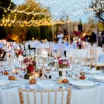 Une table élégamment dressée pour un grand événement, au cœur d'un jardin romantique sous un ciel étoilé. Les guirlandes lumineuses créent une atmosphère chaleureuse et sophistiquée, parfaite pour une célébration de mariage de luxe. Chaque détail, des fleurs délicates aux verres en cristal, reflète l'expertise de By Mademoiselle C dans l'organisation de mariages sur mesure en Provence, Côte d'Azur et Corse. Ce cadre enchante