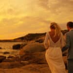 Mariage romantique au coucher du soleil sur la côte, mettant en avant un couple marchant main dans la main sur des rochers, illustrant l'expertise de By Mademoiselle C en tant que planificateur de mariage de luxe en Provence, Côte d'Azur et Corse. Ce moment unique capture l'essence d'une cérémonie grandiose dans un cadre idyllique, parfait pour une célébration sur mesure. Les couleurs chaudes du ciel ajoutent une atmosphère élégante et