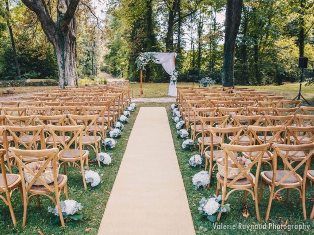 Cérémonie de mariage romantique en plein air, organisée par By Mademoiselle C, mettant en valeur un cadre élégant et naturel. Les chaises en bois délicatement disposées entourent un allée de tapis, créant une atmosphère sophistiquée pour un grand événement. Idéal pour un mariage sur mesure dans un domaine privé, ce lieu exceptionnel en Provence offre le cadre parfait pour une célébration grandiose. Les fleurs blanches ajoutent une touche de charme à cette