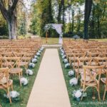 Cérémonie de mariage romantique en plein air, organisée par By Mademoiselle C, mettant en valeur un cadre élégant et naturel. Les chaises en bois délicatement disposées entourent un allée de tapis, créant une atmosphère sophistiquée pour un grand événement. Idéal pour un mariage sur mesure dans un domaine privé, ce lieu exceptionnel en Provence offre le cadre parfait pour une célébration grandiose. Les fleurs blanches ajoutent une touche de charme à cette