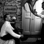 Un homme souriant s'apprête pour son mariage dans une chambre élégante, illustrant l'atmosphère romantique d'un grand événement. Cette scène, emblématique d'un mariage de luxe en Provence, Côte d'Azur et Corse, reflète l'organisation sur mesure de 'By Mademoiselle C'. La préparation minutieuse pour une cérémonie exceptionnelle souligne l'importance d'un service de planification de mariage sur mesure. L'élégance de la pièce, avec ses détails raffinés