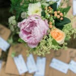 Un bouquet de fleurs délicates, comprenant des pivoines roses et des roses orangées, évoque une atmosphère romantique et élégante pour un mariage de luxe en Provence. Les éléments naturels et raffinés s'harmonisent parfaitement avec les invitations soigneusement disposées sur un fond en plein air, soulignant l'organisation sur mesure d'une cérémonie grandiose. Cette scène capture l'essence d'un événement unique, idéal pour une célébration exceptionnelle dans un cadre idyllique, que ce soit un