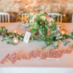 Élégante table de mariage décorée avec un centre de fleurs luxuriantes, des bougies scintillantes et une inscription "Kiss My Love", illustrant le savoir-faire de By Mademoiselle C en tant que Wedding Planner en Provence, Côte d'Azur & Corsica. Cette mise en scène romantique évoque un grand événement sur mesure, créant une atmosphère sophistiquée pour une célébration unique. Chaque détail, des arrangements floraux aux éléments de décoration,