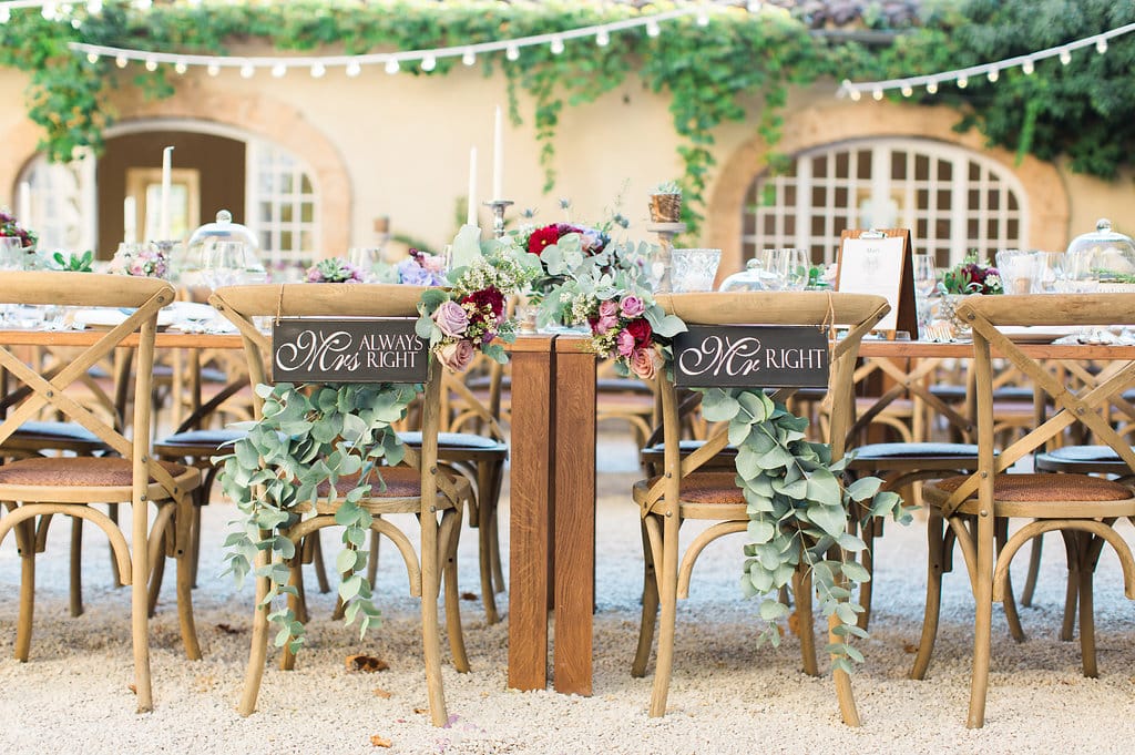 Une décoration élégante et romantique pour un mariage sur mesure dans un domaine privé en Provence, Côte d'Azur et Corse. Les chaises en bois accueillent des pancartes personnalisées "Monsieur" et "Madame", entourées de feuillage luxuriant et de fleurs délicates. Ce cadre idyllique évoque une célébration grandiose, idéale pour un événement unique. Avec des tables soigneusement dressées, cet espace extérieur crée l'atmosphère parfaite pour un mariage de