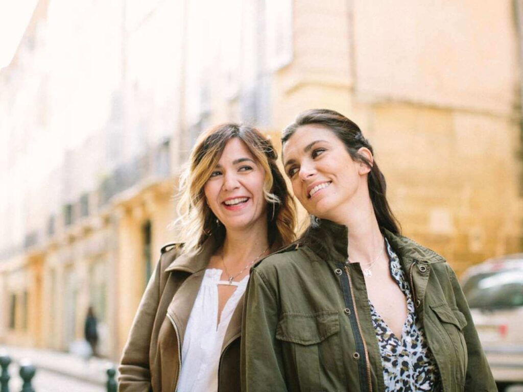 Deux femmes souriantes se tiennent dans une rue pittoresque, évoquant une atmosphère romantique et élégante, parfaite pour un mariage de luxe. 'By Mademoiselle C' excelle dans l'organisation de cérémonies sur mesure en Provence, Côte d'Azur et Corse, offrant des événements uniques dans des lieux d'exception. Créez votre rêve avec un grand célébration personnalisée.