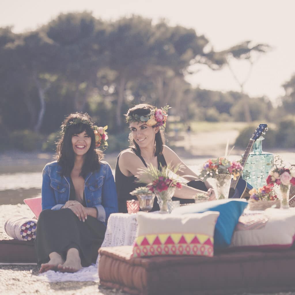 Deux femmes souriantes, entourées de fleurs et de coussins colorés, profitent d'un moment de convivialité au bord de l'eau, évoquant une atmosphère romantique idéale pour un mariage de luxe. Cette scène capture l'essence d'une célébration sur mesure dans un cadre exceptionnel, parfait pour un grand événement en Provence, Côte d'Azur et Corse. Les éléments décoratifs raffinés et l'environnement naturel créent un cadre sophistiqué, propice à un mariage de