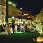 Une magnifique réception de mariage en plein air, organisée par By Mademoiselle C, se déroule dans un domaine privé en Provence. Les invités, entourés de lumières tamisées et d'une atmosphère romantique, profitent d'un grand banquet sous les étoiles. Ce luxe mariage, avec ses tables élégamment dressées, souligne la sophistication d'une célébration sur mesure. Le cadre enchanteur, agrémenté de jardins luxuriants et de pierres anciennes, en fait un lieu idéal