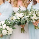 Trois demoiselles d'honneur portant des robes élégantes bleu pâle, entourées de bouquets de fleurs blanches et verdoyantes, incarnent l'atmosphère romantique d'un mariage de luxe. Cette image évoque l'organisation sur mesure d'une célébration grandiose, mettant en valeur le savoir-faire de By Mademoiselle C en tant que wedding planner en Provence, Côte d'Azur et Corse. Les éléments floraux délicats et le cadre enchanteur soul