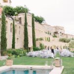 Un cadre élégant pour une célébration de mariage dans un domaine privé en Provence, où la beauté des paysages se marie à une atmosphère romantique. Cette image illustre un grand événement sur une pelouse verdoyante, entourée de cyprès majestueux et de pierres dorées, idéale pour un mariage de luxe. Les tables soigneusement dressées reflètent l'expertise de By Mademoiselle C dans l'organisation de cérémonies sur mesure. Un lieu d'exception pour un