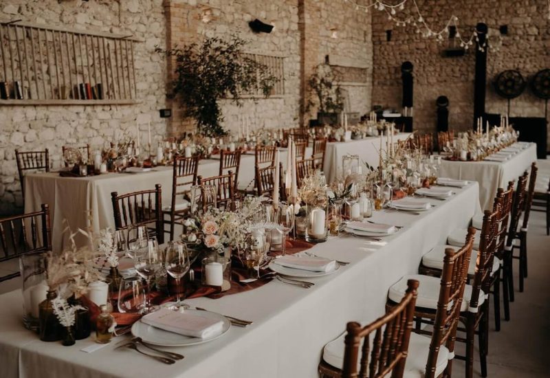 Une élégante salle de réception en pierre, décorée pour une célébration de luxe, mettant en avant le savoir-faire de By Mademoiselle C en tant que wedding planner en Provence, Côte d'Azur & Corsica. Les tables, ornées de compositions florales délicates et de bougies, créent une atmosphère romantique pour un mariage sur mesure. Ce cadre sophistiqué, avec ses murs en pierre et son ambiance chaleureuse, est idéal pour une grande cérémonie ou une