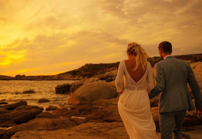 Mariage romantique au coucher du soleil sur la côte, mettant en avant un couple marchant main dans la main sur des rochers, illustrant l'expertise de By Mademoiselle C en tant que planificateur de mariage de luxe en Provence, Côte d'Azur et Corse. Ce moment unique capture l'essence d'une cérémonie grandiose dans un cadre idyllique, parfait pour une célébration sur mesure. Les couleurs chaudes du ciel ajoutent une atmosphère élégante et