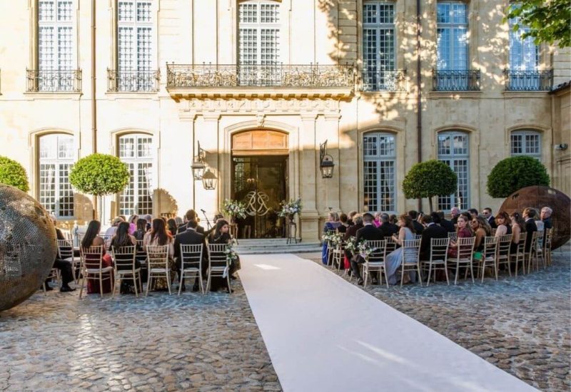 Cérémonie de mariage luxueuse en plein air dans un cadre élégant, mettant en valeur un domaine privé en Provence. L'allée blanche mène à une magnifique façade, entourée de verdure soigneusement entretenue, créant une atmosphère romantique pour une célébration sur mesure. Les invités, habillés avec raffinement, assistent à un événement unique, témoignant de l'expertise de By Mademoiselle C dans l'organisation de mariages exceptionnels. Cette scène ill