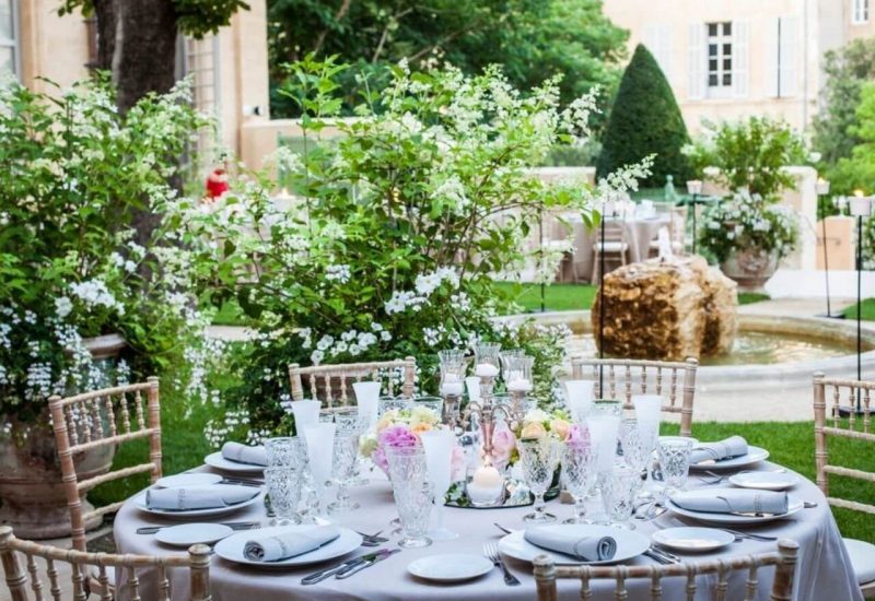 Table élégamment dressée dans un jardin luxuriant, entourée de fleurs blanches et d'arbres verdoyants, illustrant une cérémonie romantique et sophistiquée. Parfait pour un mariage de luxe, cet espace de réception dans un domaine privé en Provence évoque une célébration grandiose. Chaque détail est soigneusement conçu pour créer un événement sur mesure, offrant un cadre exceptionnel pour un mariage de rêve. L'atmosphère paisible et l'élégance des lieux font