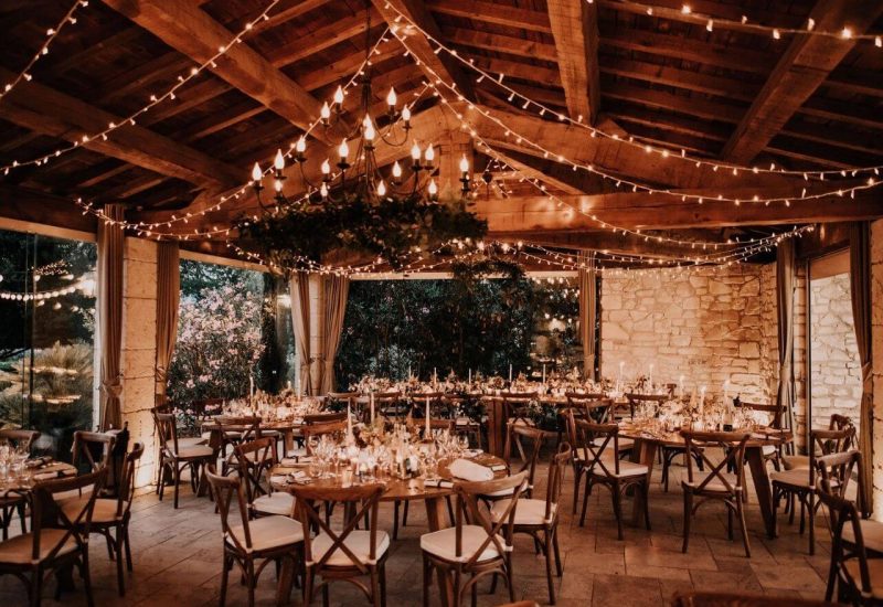 Salle élégante et romantique pour un mariage de luxe, décorée de guirlandes lumineuses et de chandeliers, offrant un cadre parfait pour une célébration grandiose. Cet espace, situé dans un domaine privé en Provence, évoque une atmosphère sophistiquée et chaleureuse, idéale pour un événement sur mesure. Les tables soigneusement dressées, entourées de verdure, créent une ambiance intime pour un mariage d'exception. Un choix parfait pour un mariage romantique