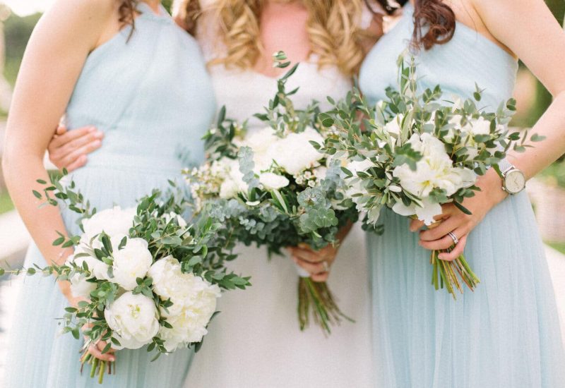 Trois demoiselles d'honneur portant des robes élégantes bleu pâle, entourées de bouquets de fleurs blanches et verdoyantes, incarnent l'atmosphère romantique d'un mariage de luxe. Cette image évoque l'organisation sur mesure d'une célébration grandiose, mettant en valeur le savoir-faire de By Mademoiselle C en tant que wedding planner en Provence, Côte d'Azur et Corse. Les éléments floraux délicats et le cadre enchanteur soul