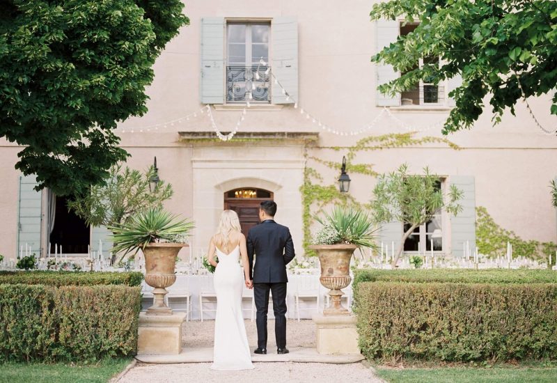 Un couple élégant se dirige vers un domaine privé, parfait pour une cérémonie grandiose. L'atmosphère romantique, entourée de verdure luxuriante et de détails raffinés, évoque un mariage sur mesure. By Mademoiselle C, votre expert en planification de mariage de luxe en Provence, Côte d'Azur et Corse, transforme vos rêves en une célébration unique et inoubliable.