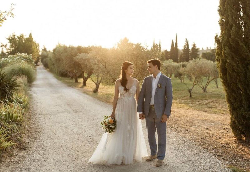 Un couple élégant se tient main dans la main sur un chemin de gravier, entouré d'oliviers en Provence, créant une atmosphère romantique et sophistiquée. Cette scène évoque la magie d'un mariage sur mesure, parfait pour une célébration grandiose dans un domaine privé. Avec des paysages pittoresques et une lumière dorée, chaque détail contribue à un événement unique et luxueux, idéal pour un rêve de mariage en Côte d'Azur et en Corse. L