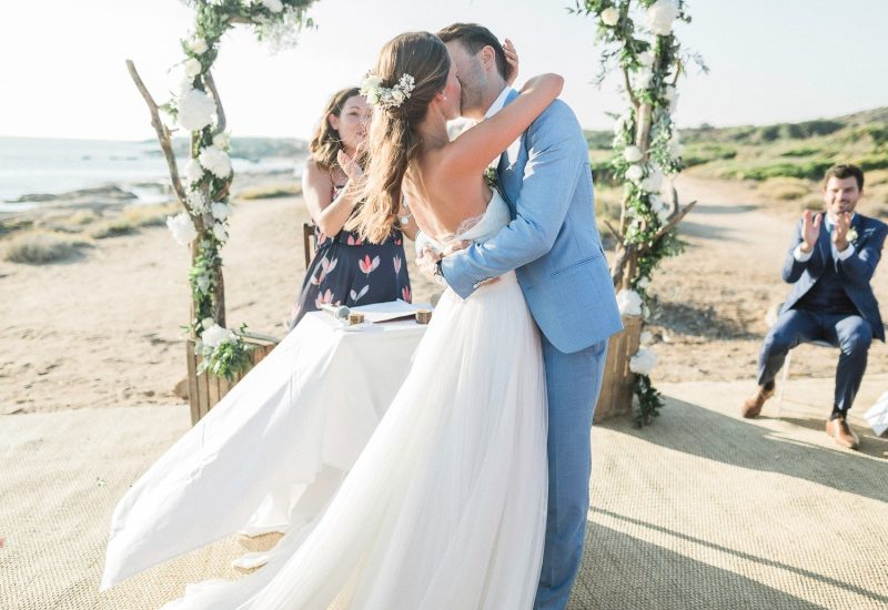 Célébration romantique d'un mariage de luxe sur une plage pittoresque, mettant en avant un couple échangeant un baiser sous un arc floral. L'atmosphère élégante et sophistiquée reflète l'expertise de By Mademoiselle C en tant que planificateur de mariage en Provence, Côte d'Azur et Corse. Cet événement sur mesure, organisé dans un cadre idyllique, évoque un rêve de mariage unique, parfait pour une grande cérémonie. Les