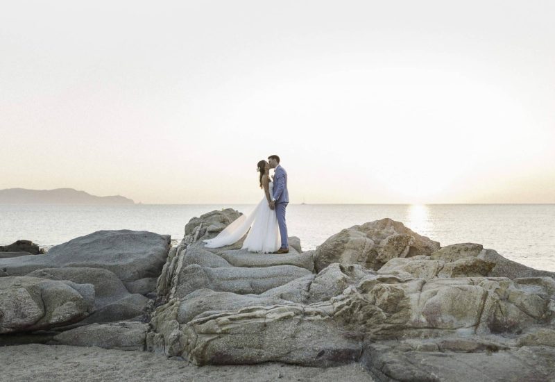 Couple romantique se tenant sur des rochers au bord de la mer, capturant l'essence d'un mariage de luxe en Provence, Côte d'Azur et Corse. Cette scène idyllique évoque une célébration grandiose, parfaite pour un événement sur mesure. L'atmosphère élégante et sophistiquée, avec le coucher de soleil en toile de fond, fait de cet endroit un domaine privé exceptionnel pour un mariage de rêve. By Mademoiselle C excelle dans l