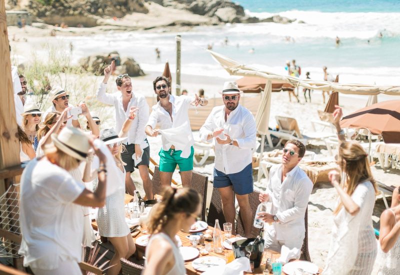 Une célébration luxueuse sur la plage en Provence, Côte d'Azur, mettant en valeur une ambiance romantique et élégante. Ce mariage sur la plage, organisé par By Mademoiselle C, incarne le rêve d'une cérémonie sur mesure, entourée par des amis joyeux. Les invités, habillés en blanc, profitent d'un cadre idyllique, avec des parasols et des chaises longues en arrière-plan, créant une atmosphère de grand événement. Chaque détail