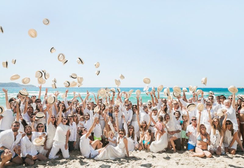 Une célébration en bord de mer réunissant des invités élégamment vêtus, illustrant un mariage romantique et une ambiance festive. Ce grand événement, organisé par 'By Mademoiselle C', incarne l'esprit d'un mariage de luxe en Provence, Côte d'Azur et Corse. Les invités, habillés de blanc, lancent des chapeaux dans les airs, créant une atmosphère joyeuse et mémorable. Ce moment époustouflant se déroule dans un