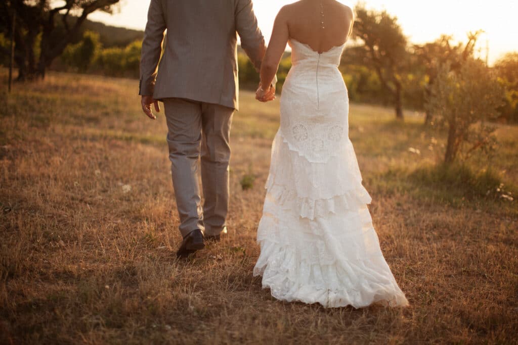 Un couple élégant marchant main dans la main dans un champ doré, symbolisant un mariage romantique et sur mesure. Ce moment capturé évoque l'atmosphère intime d'une célébration grandiose dans un domaine privé en Provence. La robe de mariée raffinée et le costume chic reflètent le style sophistiqué des événements organisés par 'By Mademoiselle C', experts en planification de mariages de luxe sur la Côte d'Azur et en Corse. Ce cadre