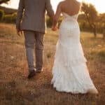 Un couple élégant marchant main dans la main dans un champ doré, symbolisant un mariage romantique et sur mesure. Ce moment capturé évoque l'atmosphère intime d'une célébration grandiose dans un domaine privé en Provence. La robe de mariée raffinée et le costume chic reflètent le style sophistiqué des événements organisés par 'By Mademoiselle C', experts en planification de mariages de luxe sur la Côte d'Azur et en Corse. Ce cadre