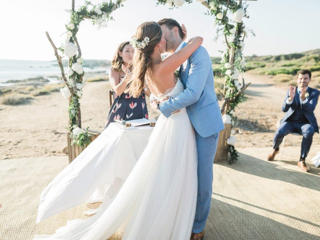 Célébration romantique d'un mariage de luxe sur une plage pittoresque, mettant en avant un couple échangeant un baiser sous un arc floral. L'atmosphère élégante et sophistiquée reflète l'expertise de By Mademoiselle C en tant que planificateur de mariage en Provence, Côte d'Azur et Corse. Cet événement sur mesure, organisé dans un cadre idyllique, évoque un rêve de mariage unique, parfait pour une grande cérémonie. Les