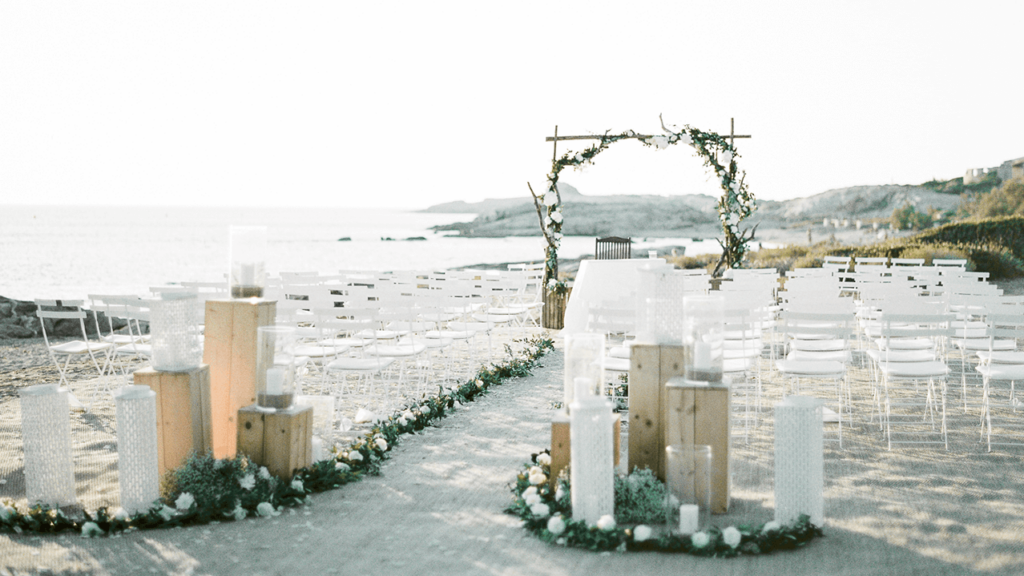 Cérémonie de mariage romantique sur la plage, mettant en avant une organisation sur mesure par By Mademoiselle C, wedding planner en Provence, Côte d'Azur & Corsica. Cette scène élégante présente des chaises blanches disposées avec soin, entourées de lanternes délicates et de fleurs, créant une atmosphère intime pour une célébration grandiose. Le cadre idyllique, avec la mer en arrière-plan, évoque un événement unique et luxueux,